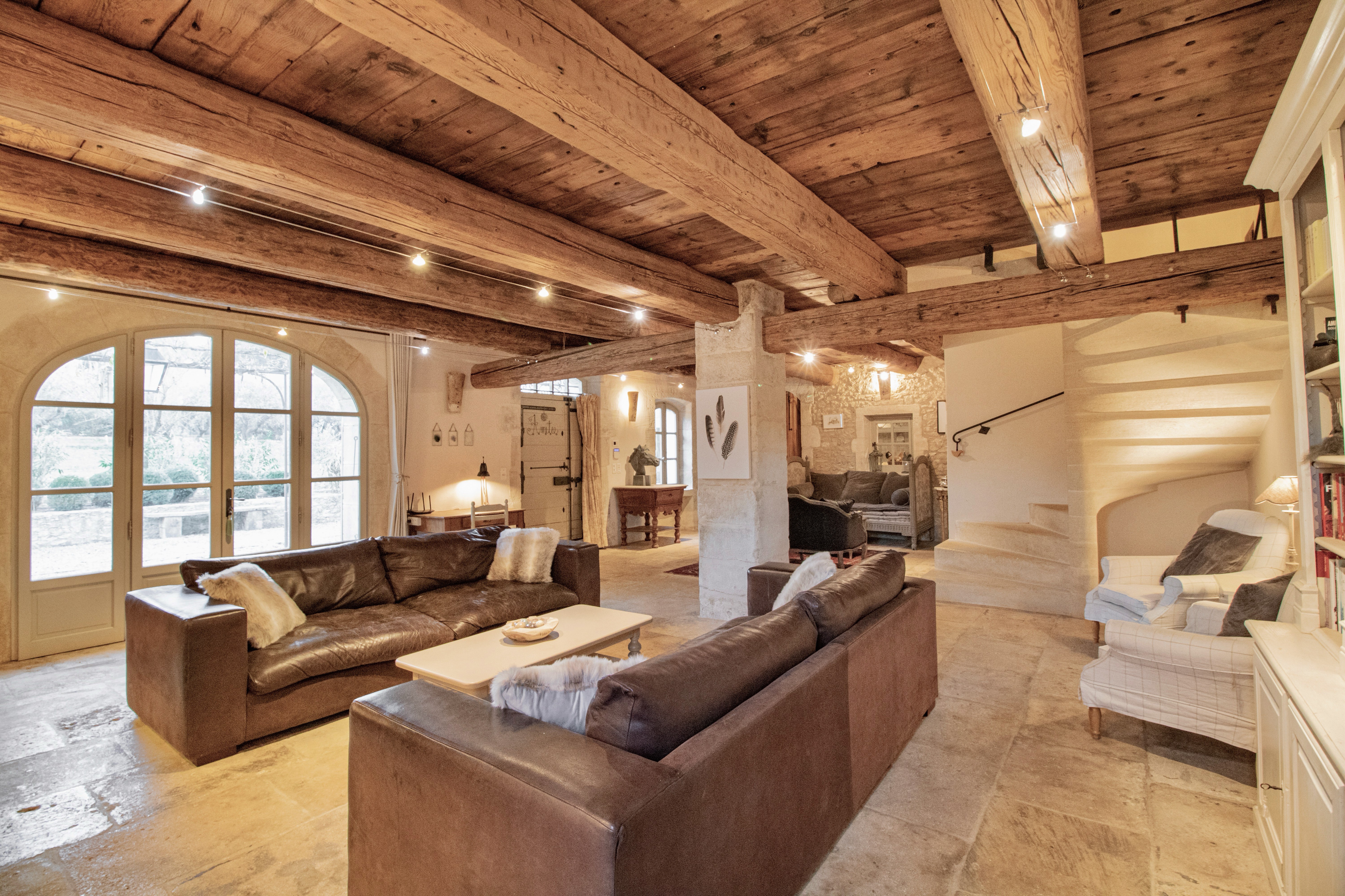 Spacious living room with exposed wooden beams, plush leather sofas, arched doors, and a staircase leading to an upper level. the room has a warm, rustic ambiance with natural light.